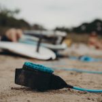 Dreamsea Costa Rica Surf Camp | backpacking, travel, hostel, surf, and yoga camp | Media Files | Lifestyle Image of Surf and Yoga Camp Resort | picture of a surf board blurred in the background with the ankle bracelet shown in focus in the foreground on a beach in Tamarindo, Costa Rica